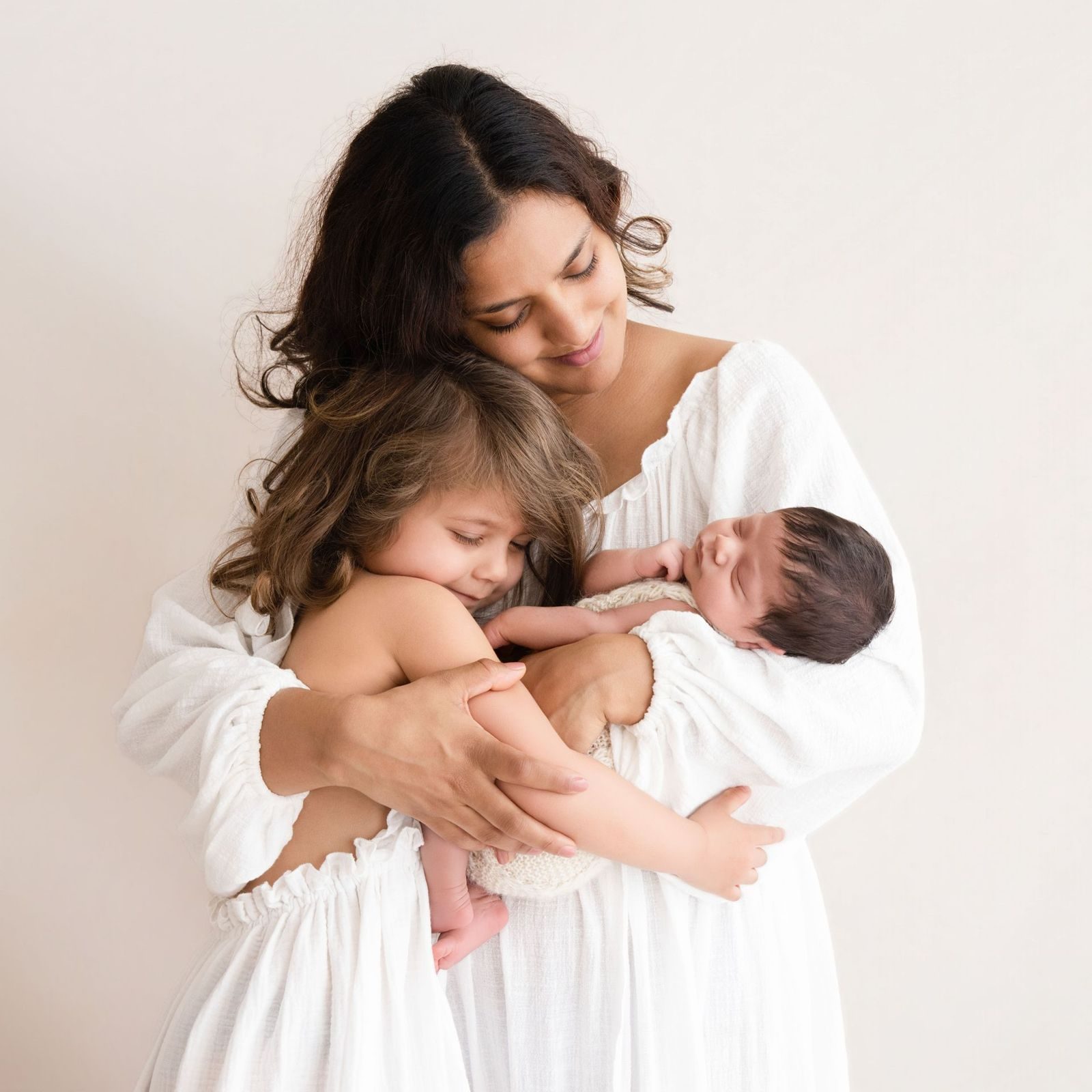 Mother hugging her toddler and newborn in white