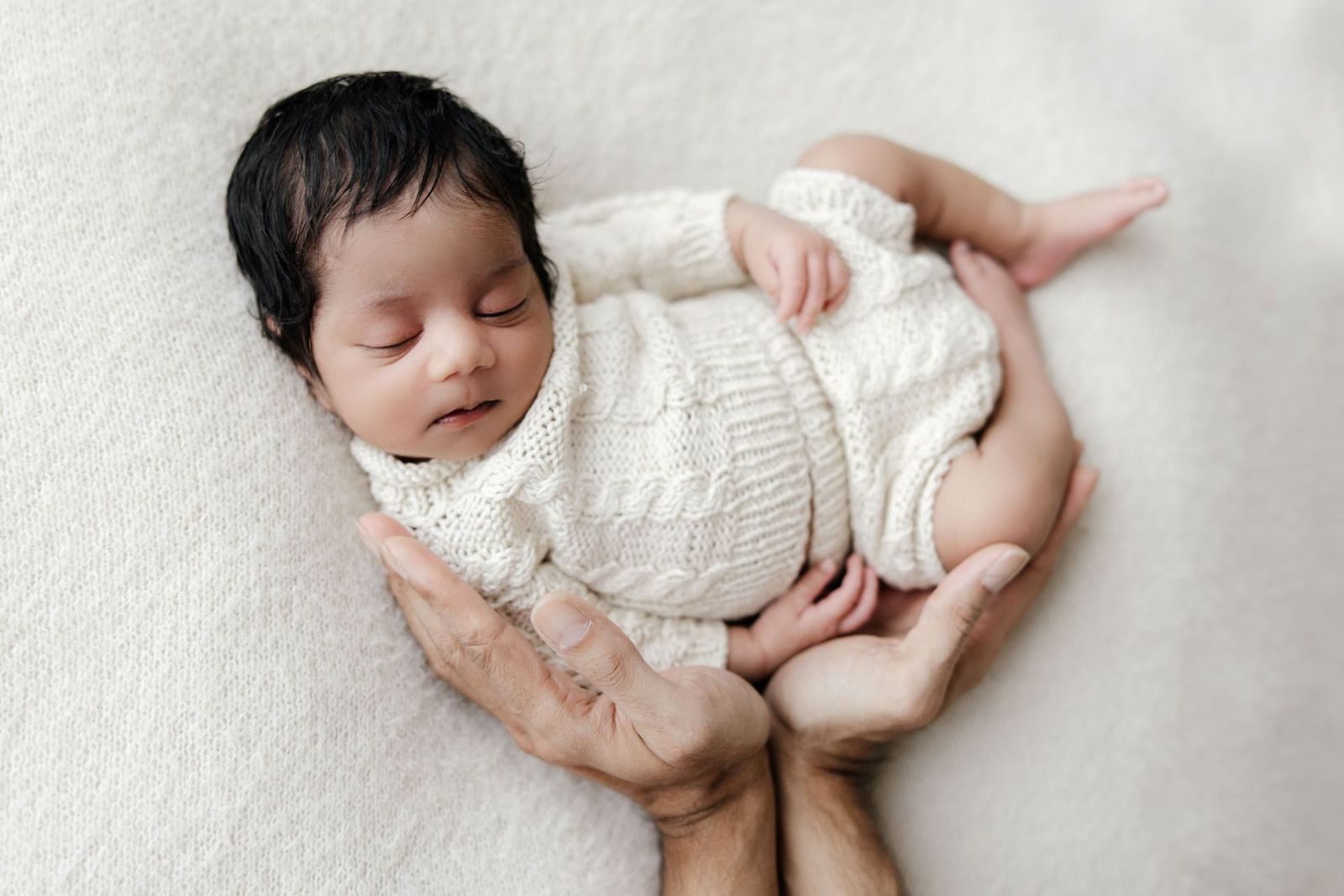 Newborn in a cable knit romper held by her father's hands